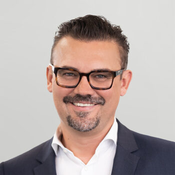 Ein Mann mit kurzem, dunklem Haar, Brille und Spitzbart lächelt, während er eine dunkle Anzugsjacke und ein weißes Hemd vor einem hellgrauen Hintergrund trägt.