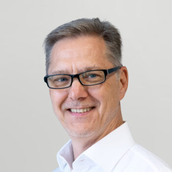 Ein Mann mittleren Alters mit kurzen grauen Haaren, schwarz umrandeter Brille und weißem Kragenhemd, lächelt vor einem schlichten hellgrauen Hintergrund.