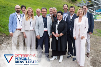 Eine Gruppe von zwölf professionell gekleideten Personen steht lächelnd im Freien auf einem Grashang neben einer Skisprungschanze. In der unteren linken Ecke ist das Logo
