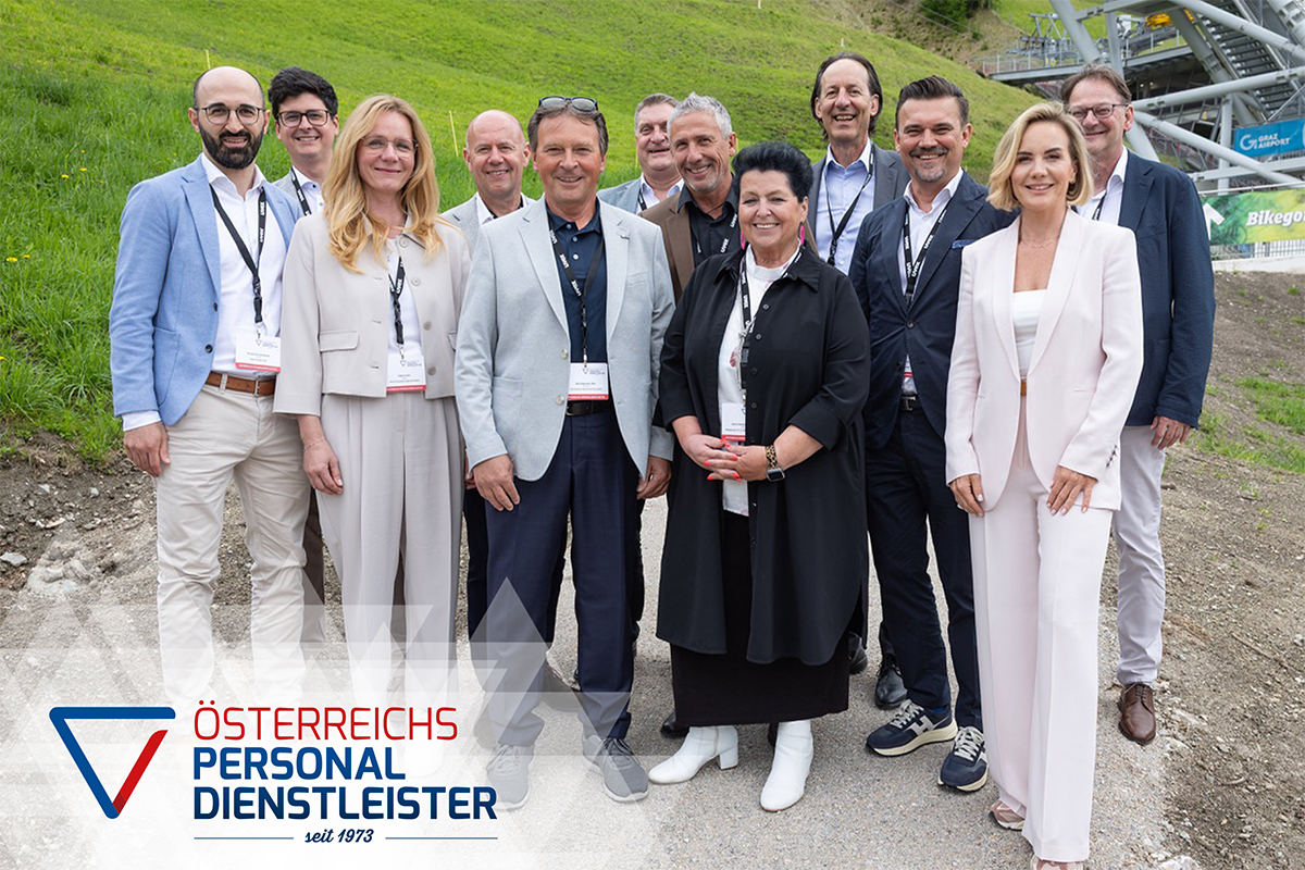 Eine Gruppe von zwölf professionell gekleideten Personen steht lächelnd im Freien auf einem Grashang neben einer Skisprungschanze. In der unteren linken Ecke ist das Logo "Österreichs Personaldienstleister" zu sehen.