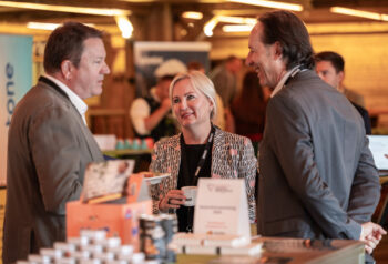 Drei professionell gekleidete Personen stehen auf einer Konferenz oder einem Networking-Event und unterhalten sich, l&auml;chelnd und mit Tassen in der Hand, auf dem Tisch vor ihnen liegen Informationsmaterialien und Firmenaush&auml;nge.