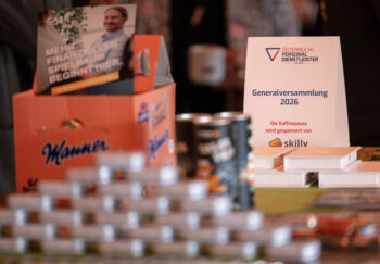 Nahaufnahme eines Tisches mit Snacks, Kaffeedosen und einem Schild mit der Aufschrift Generalversammlung 2026 by &Ouml;sterreichs Personaldienstleister, auf dem steht, dass die Kaffeepause von skillv gesponsert wird. Die Menschen im Hintergrund sind unscharf.