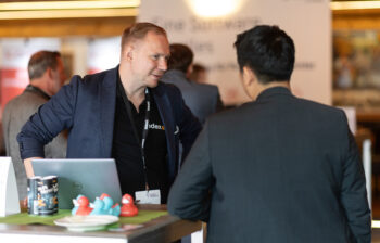 Zwei M&auml;nner unterhalten sich an einem Konferenzstand; einer blickt in die Kamera und l&auml;chelt, w&auml;hrend er hinter einem Laptop und Werbeartikeln, darunter Gummienten, steht. Andere Personen und Banner sind im Hintergrund unscharf.