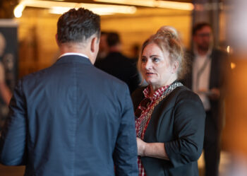Eine Frau mit blondem Haar in einem Dutt unterh&auml;lt sich mit einem Mann im Anzug bei einer Indoor-Veranstaltung. Der Hintergrund ist leicht unscharf und zeigt andere Menschen und warmes Licht.