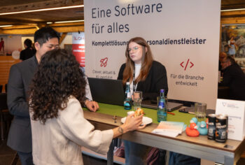 Drei Personen unterhalten sich am Stand eines Softwareunternehmens auf einer Veranstaltung. Eine Frau steht mit einem Laptop hinter der Theke, w&auml;hrend zwei weitere Personen davor stehen. Auf dem Tresen stehen Werbematerialien und Getr&auml;nke. Auf dem Stand steht ein deutscher Text.