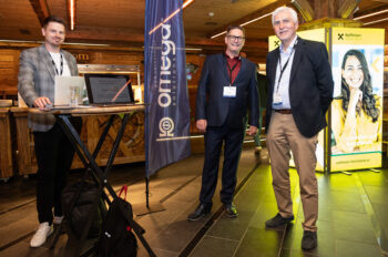 Drei M&auml;nner in Gesch&auml;ftskleidung stehen in einem Innenraum in der N&auml;he eines Laptops und von Bannern f&uuml;r Omega und die Raiffeisen Bank. Sie l&auml;cheln und scheinen an einer professionellen Netzwerk- oder Konferenzveranstaltung teilzunehmen.