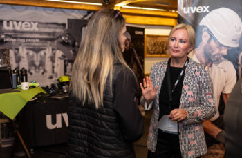 Eine blonde Frau in einem gemusterten Blazer unterh&auml;lt sich mit einer anderen Frau an einem Veranstaltungsstand, an dem im Hintergrund UVEX-Branding, Sicherheitsausr&uuml;stung und Poster zu sehen sind.