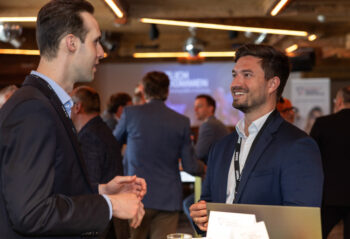 Zwei M&auml;nner in Gesch&auml;ftskleidung unterhalten sich bei einer Networking-Veranstaltung und l&auml;cheln dabei. Andere Personen in Anz&uuml;gen unterhalten sich im Hintergrund, und die Atmosph&auml;re wirkt professionell und freundlich.
