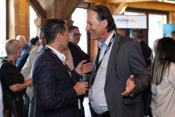 Zwei M&auml;nner in Gesch&auml;ftskleidung l&auml;cheln und unterhalten sich bei einer Networking-Veranstaltung, wobei sie Schl&uuml;sselb&auml;nder tragen. Andere Leute mischen sich im Hintergrund in einem warm beleuchteten Raum mit Holzbalken.