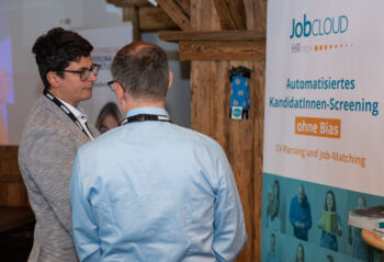 Zwei M&auml;nner stehen und unterhalten sich in der N&auml;he eines Banners mit der Aufschrift JobCloud HR Tech: Automatisiertes KandidatInnen-Screening ohne Bias, CV-Parsing und Job-Matching auf Deutsch. Die Szene findet in einem Innenraum statt, in dem Holzbalken zu sehen sind.
