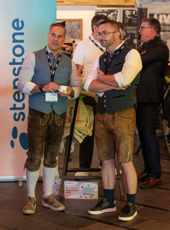 Zwei M&auml;nner in bayerischer Tracht unterhalten sich auf einer Veranstaltung neben einer blauen Stemstone-Fahne. Beide tragen Namensschilder, und im Hintergrund unterhalten sich weitere Personen.