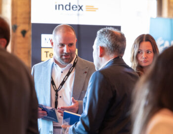 Ein Mann in einem grauen Anzug spricht mit einem anderen Mann bei einer Networking-Veranstaltung und h&auml;lt eine Brosch&uuml;re in der Hand. Eine Frau steht in der N&auml;he und schaut zu ihnen hin. Im Hintergrund ist ein Banner mit dem Wort Index zu sehen.