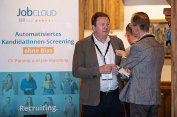 Zwei M&auml;nner in Gesch&auml;ftskleidung unterhalten sich vor einem JobCloud HR Tech-Banner, das auf einer Fachveranstaltung f&uuml;r ein automatisches Bewerberscreening ohne Vorurteile wirbt. Ein Mann h&auml;lt ein Smartphone in der Hand, w&auml;hrend sie sich unterhalten.