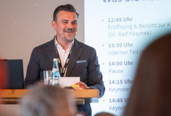 Ein Mann im Anzug mit einem Headset-Mikrofon steht l&auml;chelnd an einem Podium, vor ihm ein Glas Wasser und Papiere. An der Wand hinter ihm ist ein Pr&auml;sentationsplan in deutscher Sprache projiziert.