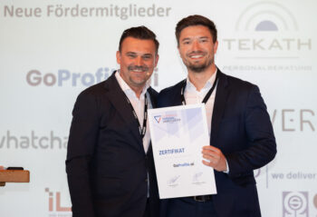 Zwei M&auml;nner in Anz&uuml;gen l&auml;cheln in die Kamera. Einer h&auml;lt eine Urkunde mit der Aufschrift ZERTIFIKAT in der Hand. Hinter ihnen ist ein Bildschirm mit Logos und Text in deutscher Sprache zu sehen.