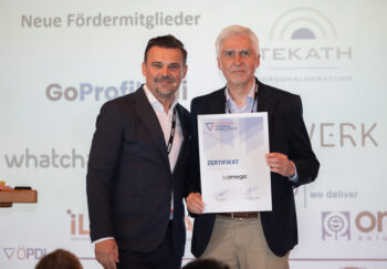 Zwei M&auml;nner in Gesch&auml;ftskleidung stehen bei einer Veranstaltung zusammen. Einer h&auml;lt ein Zertifikat mit der Aufschrift Zertifikat. Auf der Leinwand hinter ihnen werden Logos und Texte von verschiedenen Unternehmen projiziert.