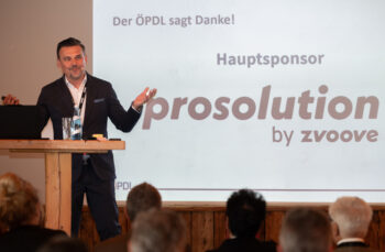 Ein Mann im Anzug steht auf einem Podium und spricht mit Gesten zu einem Publikum. Hinter ihm ist ein Bildschirm mit dem Text Der &Ouml;PDL sagt Danke! und Hauptsponsor prosolution by zvoove zu sehen.