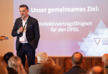 Ein Mann im Anzug spricht zu einem Publikum vor einer Pr&auml;sentationsfolie in deutscher Sprache, auf der zu lesen ist: Unser gemeinsames Ziel: Kollektivvertragsf&auml;higkeit f&uuml;r den &Ouml;PDL.
