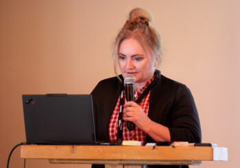 Eine Frau mit blondem Dutt, rotkariertem Hemd und schwarzer Jacke spricht in ein Mikrofon, w&auml;hrend sie an einem Rednerpult steht, auf dem ein aufgeklappter Laptop und einige kleine Gegenst&auml;nde liegen.