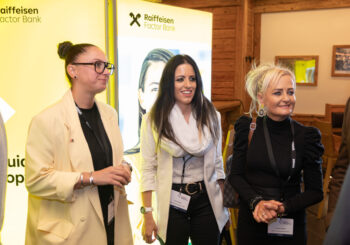Drei Frauen stehen zusammen und l&auml;cheln bei einem Indoor-Event, sie tragen Namensschilder. Hinter ihnen befinden sich ein helles Display der Raiffeisen-Factor-Bank und Holzw&auml;nde, die auf ein berufliches Treffen oder eine Netzwerkveranstaltung hindeuten.
