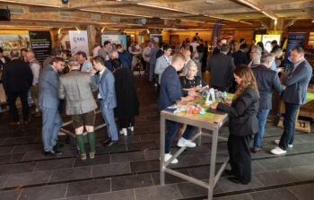 Eine Gruppe von Personen in Gesch&auml;ftskleidung unterh&auml;lt sich bei einer Indoor-Veranstaltung mit Firmenbannern, die um Stehtische in einem rustikalen Holzgeb&auml;ude stehen. Auf den Tischen sind Getr&auml;nke und Werbematerialien zu sehen.