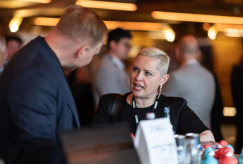 Zwei Personen in Gesch&auml;ftskleidung unterhalten sich bei einem Networking-Event. Die Frau mit kurzen blonden Haaren und gro&szlig;en Ohrringen steht dem Mann gegen&uuml;ber und gestikuliert mit ihrer Hand. Im Hintergrund sind unscharfe Menschen und warmes Licht zu sehen.