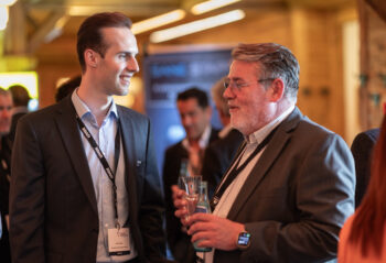 Zwei M&auml;nner in Gesch&auml;ftskleidung unterhalten sich und l&auml;cheln bei einem beruflichen Networking-Event. Ein Mann h&auml;lt ein Glas Wasser, und beide tragen Namensschilder an Schl&uuml;sselb&auml;ndern. Andere Personen sind im unscharfen Hintergrund zu sehen.