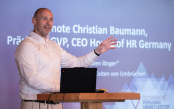 Ein Mann im wei&szlig;en Hemd steht an einem Podium und gestikuliert mit der rechten Hand, w&auml;hrend er pr&auml;sentiert. Hinter ihm zeigt ein Bildschirm seinen Namen, Christian Baumann, und seinen Titel als CEO von HR Deutschland.