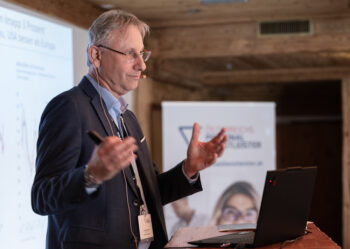 Ein Mann im Anzug steht an einem Podium und h&auml;lt mit seinen H&auml;nden eine Pr&auml;sentation. Vor ihm ist ein Laptop aufgeklappt, und im Hintergrund sind eine Projektionsfl&auml;che und ein verschwommenes Banner zu sehen.