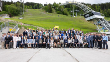 Eine gro&szlig;e Gruppe von Personen in Gesch&auml;ftskleidung posiert f&uuml;r ein Gruppenfoto im Freien vor einem grasbewachsenen H&uuml;gel, mit Skiliftanlagen und B&auml;umen im Hintergrund.
