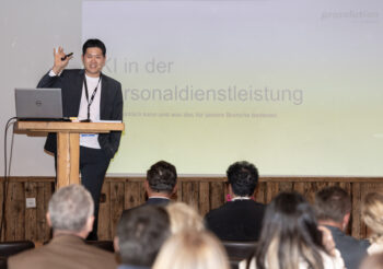 Ein Mann im Anzug steht auf einem Podium und h&auml;lt einen Vortrag vor einem Publikum. Er gestikuliert mit einer Hand und l&auml;chelt. Hinter ihm ist eine projizierte Folie mit deutschem Text zu sehen.