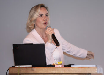 Eine Frau mit kurzen blonden Haaren in einem hellen Blazer spricht leidenschaftlich in ein Mikrofon, w&auml;hrend sie an einem Holzpodium steht, auf dem ein Laptop und Papiere liegen. Ihr Arm ist zur Seite ausgestreckt.