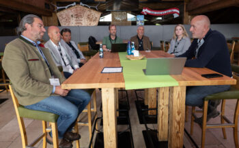 Sieben Personen, sechs M&auml;nner und eine Frau, sitzen um einen Holztisch in einem rustikalen Raum und diskutieren. Laptops, Getr&auml;nke und Papiere liegen auf dem Tisch. Die Atmosph&auml;re scheint professionell und kooperativ zu sein.
