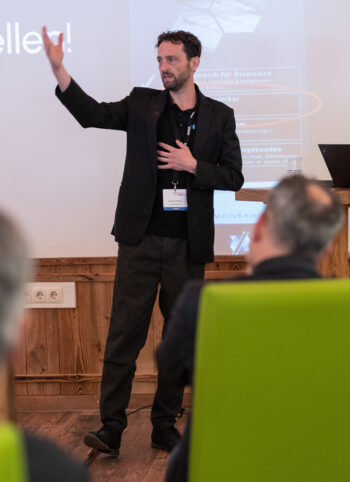 Ein Mann in einem schwarzen Anzug steht und gestikuliert, w&auml;hrend er zu einem Publikum in einem Konferenzraum spricht. Er tr&auml;gt ein Schl&uuml;sselband und ein Namensschild. Eine Pr&auml;sentation wird auf den Bildschirm hinter ihm projiziert.