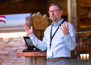 Ein Mann mit Brille und hellblauem Hemd spricht w&auml;hrend einer Pr&auml;sentation leidenschaftlich mit erhobenen H&auml;nden. Er steht an einem Holzpodium mit einem Laptop und tr&auml;gt ein Schl&uuml;sselband mit einem Namensschild.