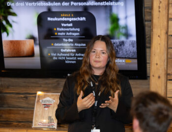Eine Frau in einem schwarzen Blazer pr&auml;sentiert vor einem Bildschirm, auf dem ein deutscher Text &uuml;ber den Verkauf von Personaldienstleistungen angezeigt wird. Sie steht in einem Innenraum an holzget&auml;felten W&auml;nden und gestikuliert mit den H&auml;nden, w&auml;hrend sie spricht.