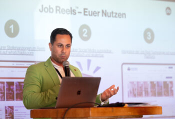 Ein Mann in einem gr&uuml;nen Blazer spricht an einem Podium mit einem Laptop und pr&auml;sentiert Folien &uuml;ber Job Reels - Euer Nutzen auf Deutsch. Auf der Folie hinter ihm sind nummerierte Punkte und Screenshots von Websites abgebildet.