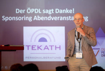 Ein Mann steht mit einem Mikrofon in der Hand vor einem Bildschirm, auf dem die Worte Der &Ouml;PDL sagt Danke! Sponsoring Abendveranstaltung und ein TEKATH-Logo. Er tr&auml;gt eine Brille, ein Schl&uuml;sselband und eine beige Jacke &uuml;ber einem blauen Hemd.