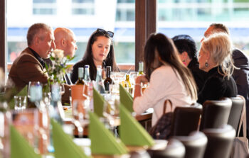 Eine Gruppe von Menschen sitzt um einen Tisch in einem Restaurant und unterh&auml;lt sich. Der Tisch ist mit gr&uuml;nen Servietten, Gl&auml;sern, Flaschen und Blumen gedeckt. Sonnenlicht str&ouml;mt durch die Fenster hinter ihnen.