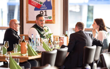 Vier Erwachsene sitzen um einen mit Getr&auml;nken, Weinflaschen und gr&uuml;nen Servietten gedeckten Restauranttisch und unterhalten sich. Sonnenlicht str&ouml;mt herein, und an der Wand hinter ihnen h&auml;ngt ein gerahmtes Trikot.