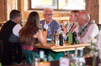 Eine Gruppe von f&uuml;nf Personen sitzt an einem Restauranttisch und unterh&auml;lt sich. Auf dem Tisch stehen Flaschen mit Wasser und Getr&auml;nken, und die Atmosph&auml;re ist warm und gem&uuml;tlich, mit Holzw&auml;nden im Hintergrund.