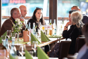 Eine Gruppe von Menschen sitzt um einen mit Getr&auml;nken, Gl&auml;sern und gefalteten gr&uuml;nen Servietten gedeckten Restauranttisch und unterh&auml;lt sich. Das Sonnenlicht f&auml;llt durch gro&szlig;e Fenster im Hintergrund.