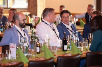 Drei M&auml;nner in Gesch&auml;ftskleidung sitzen an einem Tisch mit Getr&auml;nken und gr&uuml;nen Servietten und unterhalten sich w&auml;hrend einer formellen Veranstaltung in einem warm beleuchteten Raum. Mehrere andere Teilnehmer sind im Hintergrund zu sehen.