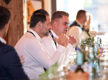 Zwei M&auml;nner in wei&szlig;en Hemden sitzen nebeneinander an einem mit Gl&auml;sern, Flaschen und Blumen gedeckten Tisch und unterhalten sich bei einer hellen Veranstaltung oder einem Treffen in einem Haus.