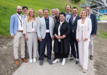 Eine Gruppe von elf professionell gekleideten Personen steht l&auml;chelnd im Freien auf einem Weg, mit einem grasbewachsenen H&uuml;gel und einer Skisprungschanze im Hintergrund.