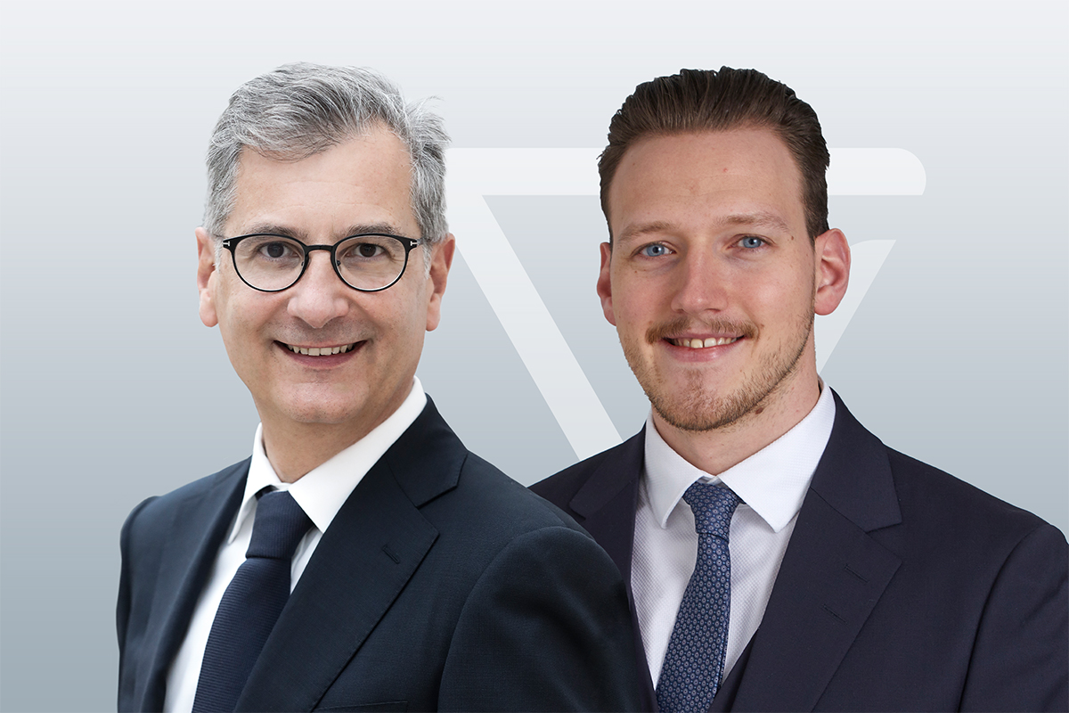 Zwei Männer in Anzug und Krawatte lächeln in die Kamera. Der Mann links hat graues Haar, trägt eine Brille und einen dunklen Anzug. Der Mann rechts hat hellbraunes Haar und trägt ebenfalls einen dunklen Anzug. Der Hintergrund ist hellgrau.