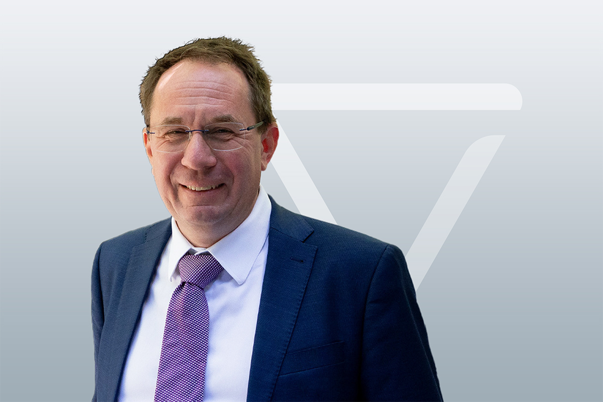 Ein Mann mit Brille, in einem blauen Anzug und einer lila gemusterten Krawatte lächelt in die Kamera. Der Hintergrund ist hellgrau und weist ein dezentes, abstraktes geometrisches Muster auf.