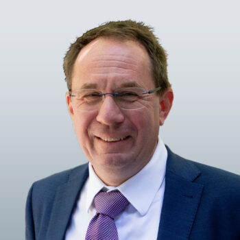 Ein Mann mittleren Alters mit Brille, in einem blauen Anzug, einem wei&szlig;en Hemd und einer violett gemusterten Krawatte, der vor einem hellgrauen Hintergrund l&auml;chelt.