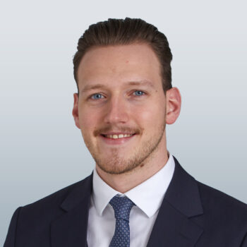 Ein Mann mit kurzen braunen Haaren und einem gepflegten Bart, der einen dunklen Anzug, ein wei&szlig;es Hemd und eine gemusterte Krawatte tr&auml;gt, l&auml;chelt vor einem hellgrauen Hintergrund in die Kamera.