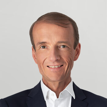 Ein Mann in einem dunklen Sakko und einem wei&szlig;en Hemd l&auml;chelt vor einem schlichten hellgrauen Hintergrund in die Kamera.